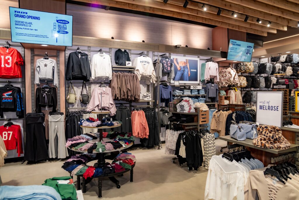 A clothing store interior with racks of hanging shirts, jackets, and folded clothes. Signs advertise a grand opening and sale. The store is well-lit with wooden accents on the ceiling and displays various apparel and accessories.