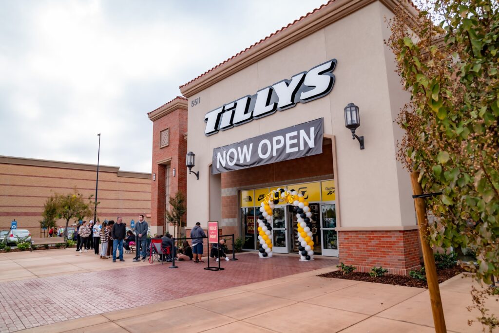 A Tillys store with a "Now Open" sign above the entrance. A small crowd of people, some with folding chairs, are lined up outside. Black and white balloon columns flank the doorway. The building has a brick exterior and a clear sky.