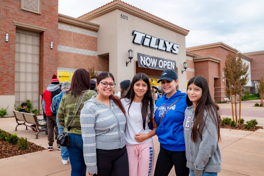 A group of four people stand together smiling in front of a Tillys store with a "Now Open" sign. The store entrance is surrounded by brick and beige walls, and people are in line outside the store.