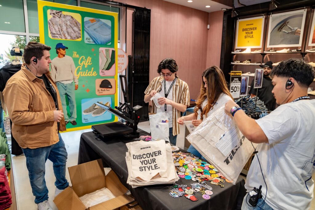 Four people are in a store setting, working on a custom clothing project. One person operates a heat press for a shirt labeled "Discover Your Style." A large poster and clothing items are displayed in the background.