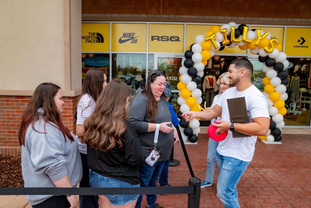 A man hands a card to a woman in a small group outside a Tilly's store. The group stands near a black and yellow balloon arch. The store has logos for The North Face, Nike, and Converse visible.