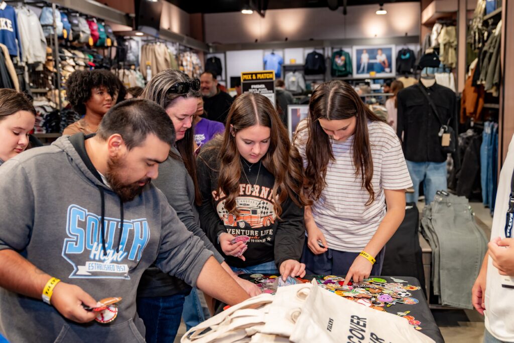 A group of people gathered around a table in a clothing store, looking at colorful pins. The store is filled with various apparel on racks and shelves. The atmosphere appears lively and interactive.