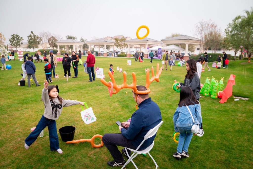 A lively outdoor event at a park features families enjoying activities. A child tosses a ring onto inflatable antlers worn by a seated person. Others engage in various games in the background, surrounded by green grass and trees.