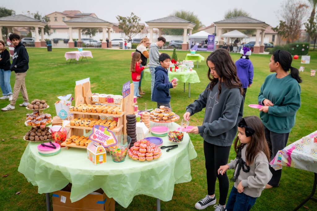 People are gathered at an outdoor picnic event with tables displaying a variety of treats, including donuts and cupcakes. A woman and child are selecting snacks from the table while others socialize in the background. It’s a cloudy day in a park setting.