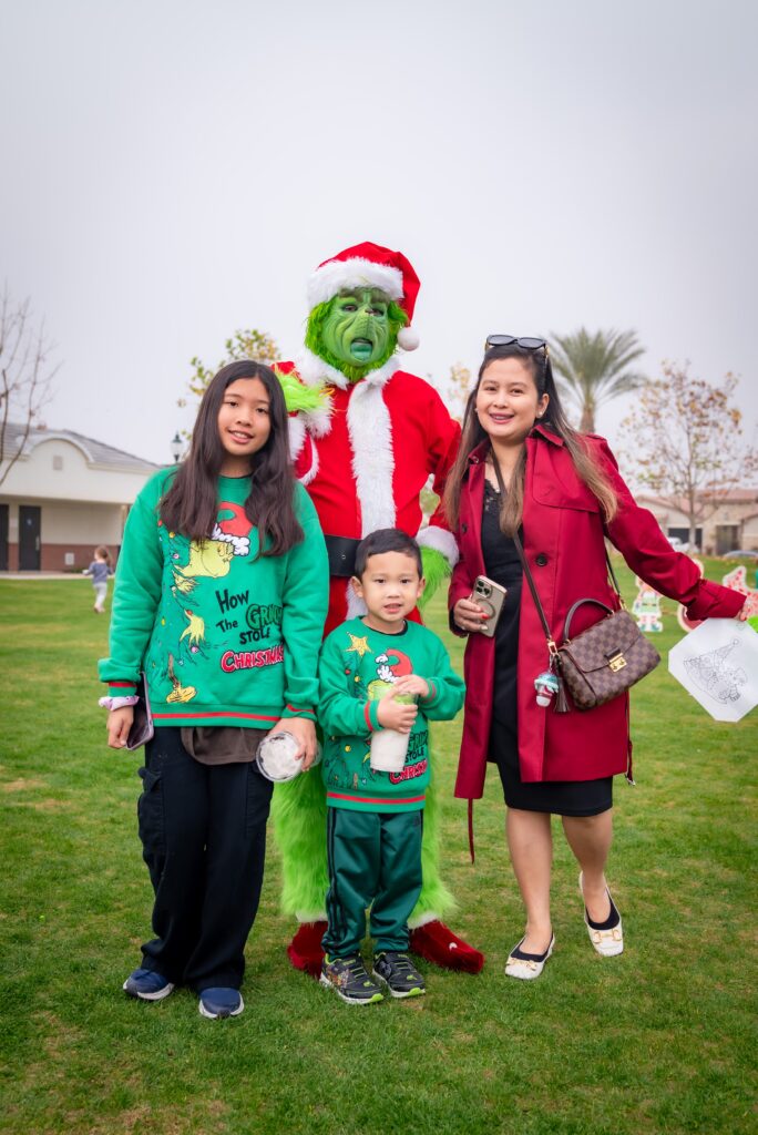 A family poses outdoors with a person dressed as the Grinch in a Santa suit. The children wear festive sweaters, and the adult is in a red coat. They stand on green grass with trees and a building in the background.