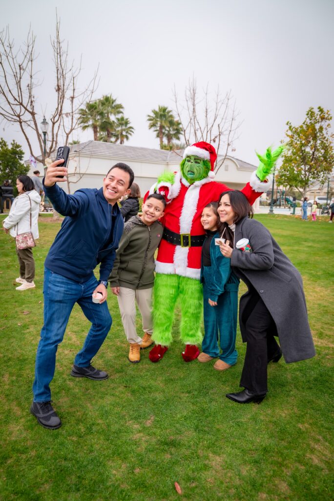 A group of people smiling and taking a selfie with someone dressed as the Grinch in a Santa outfit. They are standing on a grassy lawn with trees and buildings in the background.