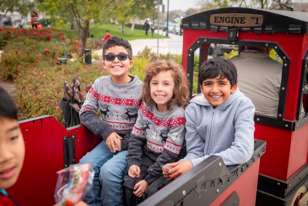 Three children smile while sitting in a miniature train car with "Engine No. 1" sign. Two wear matching holiday sweaters with reindeer patterns, and one wears a gray hoodie. They're surrounded by a park setting with trees and people in the background.