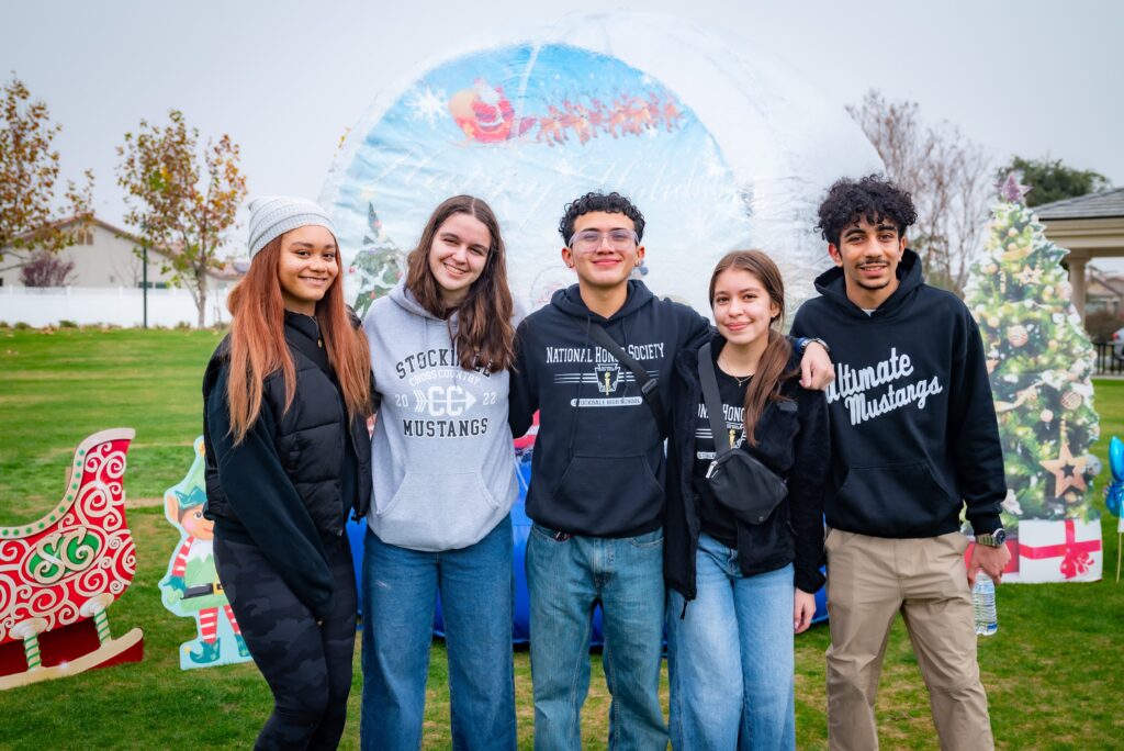 A group of five young people stand together smiling in front of a festive backdrop featuring a sleigh and Christmas tree. They are dressed casually, and the scene suggests a holiday event in a park.