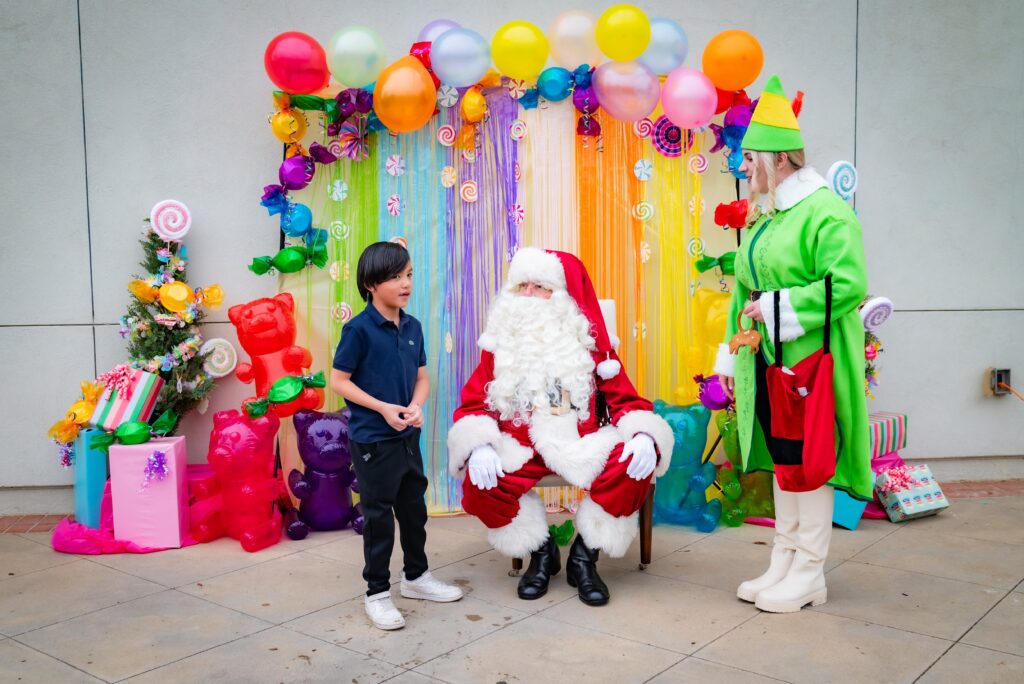 A child stands in front of a seated Santa Claus, with a colorful backdrop of balloons and ribbons. An elf in a green costume stands nearby. Brightly wrapped gifts and festive decorations surround them.