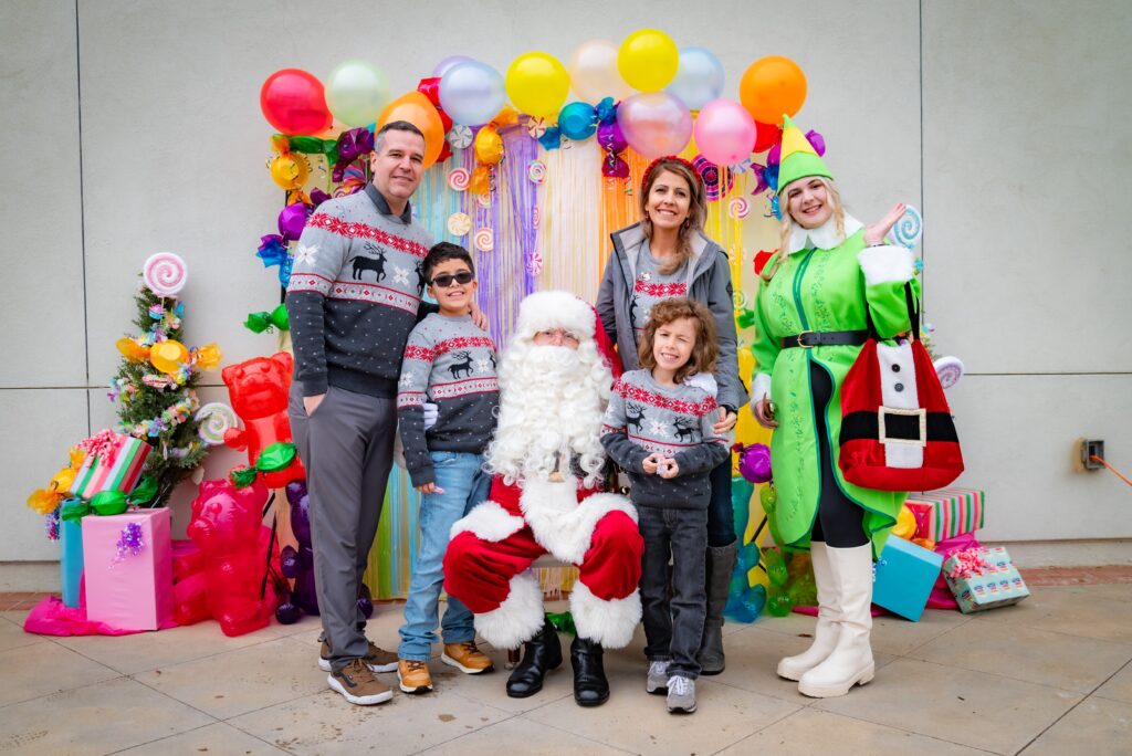 A family poses for a festive photo with Santa Claus. Two adults and two children wear matching Christmas sweaters. A colorfully dressed character stands nearby. Decorative balloons and wrapped gifts add to the holiday setting.