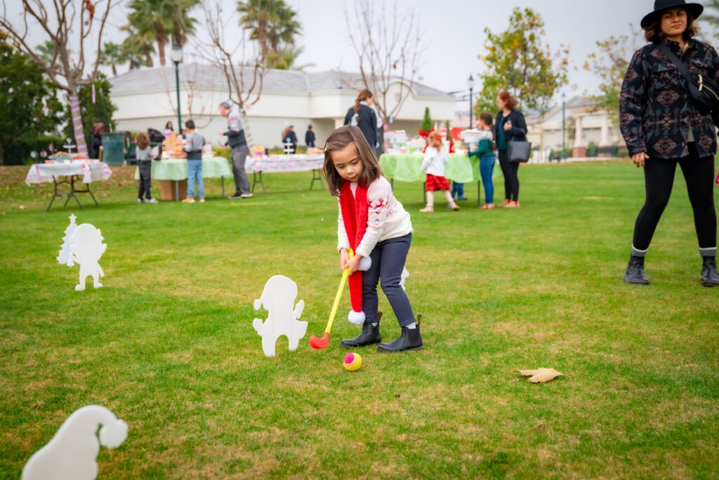 A young child in a red scarf and sweater plays mini-golf on a grassy field. People are gathered in the background with tables and holiday decorations. Trees and houses are visible in the distance.