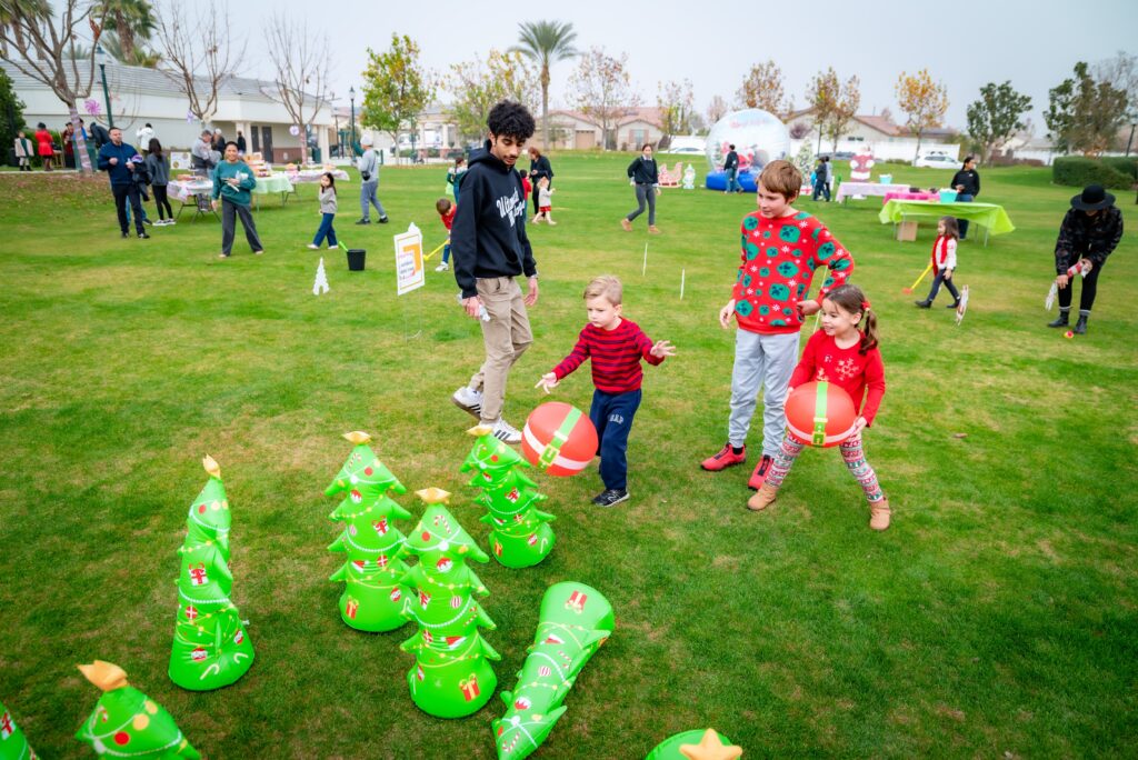 Children play a game with inflatable green Christmas trees and red balls on a grassy field. Adults and children participate in activities in the background, with a festive atmosphere and trees partially visible.