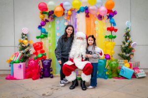 A festive scene shows two girls posing with Santa Claus in front of a colorful backdrop. The setting includes vibrant balloons, candy decorations, and wrapped gifts, creating a joyful holiday atmosphere.