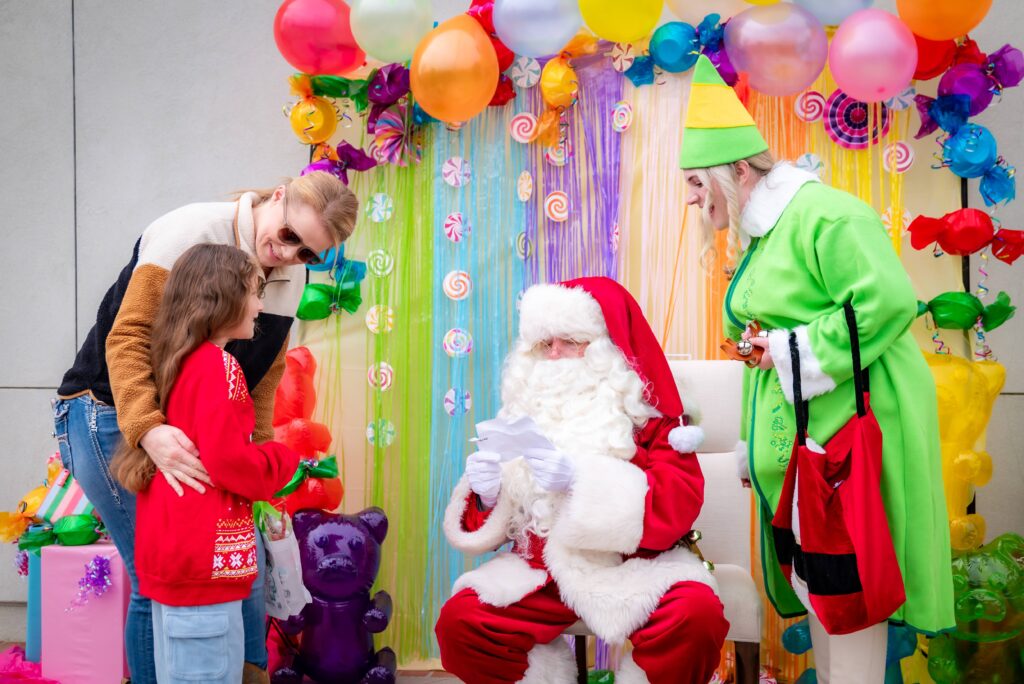 A child in a red sweater talks to a seated Santa Claus, surrounded by colorful decorations and balloons. An elf in a green outfit stands nearby, and a woman leans in towards the child, smiling. The setting is festive and bright.