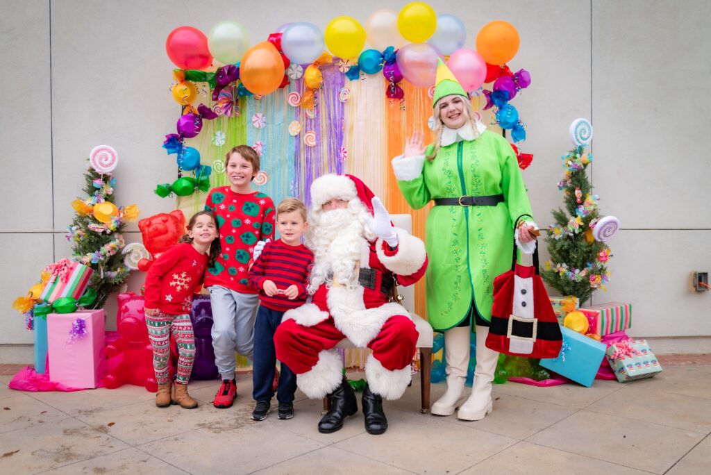 A festive scene with three children and Santa Claus posing in front of colorful decorations and balloons. An elf, wearing a green costume, stands beside them. Gift boxes and small decorated trees surround the group, adding to the holiday atmosphere.