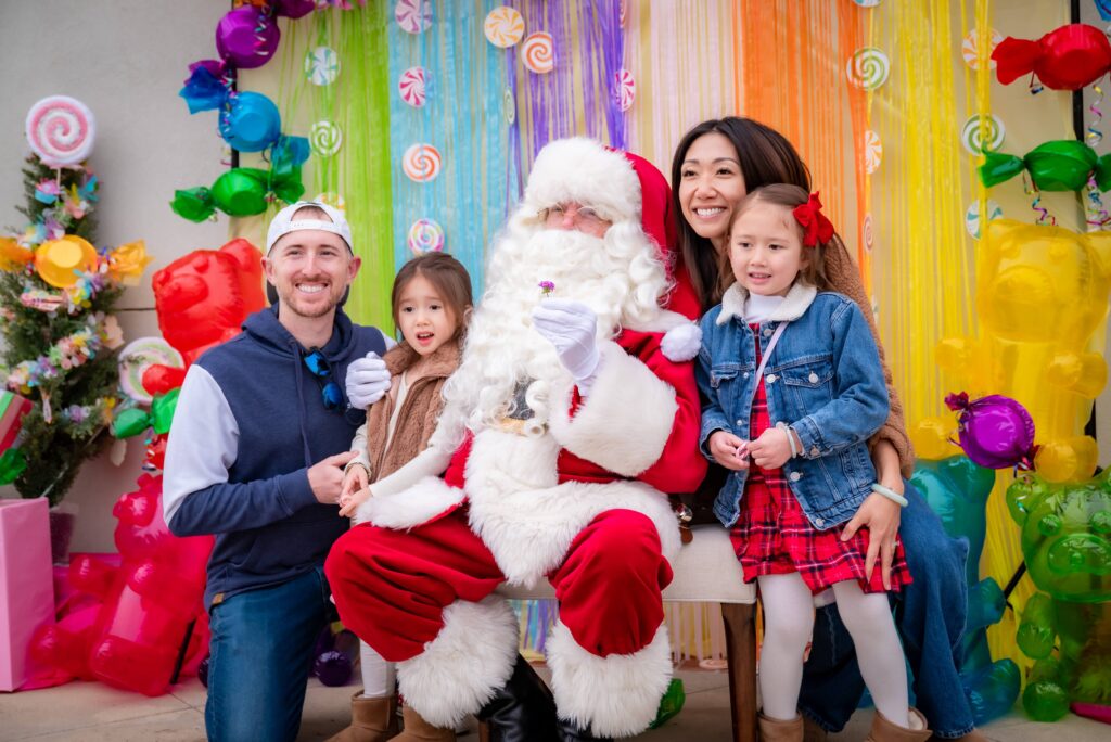 A family poses with Santa Claus in front of a colorful, candy-themed backdrop. Two adults kneel beside Santa, while two young children sit on his lap, smiling. Bright decorations and gift boxes surround them.