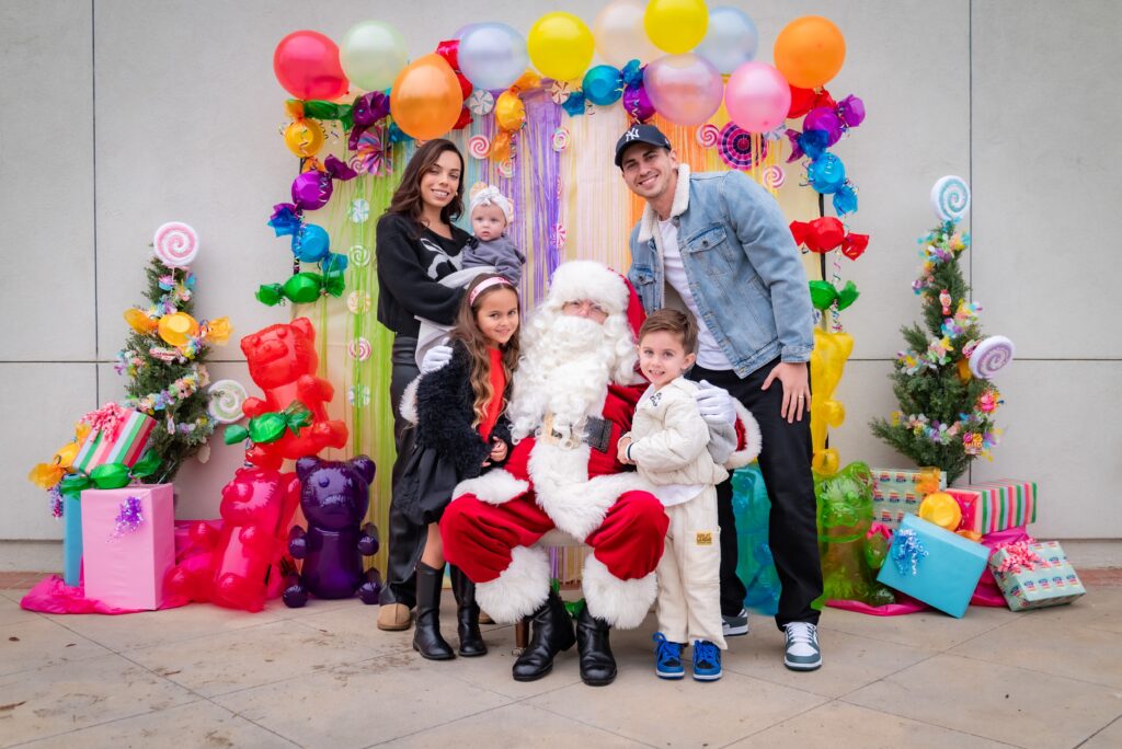 A family poses with Santa Claus in a festive setting. The background features colorful balloons and lollipop decorations, with two small decorated Christmas trees. Gift boxes are placed on either side, adding to the holiday theme.