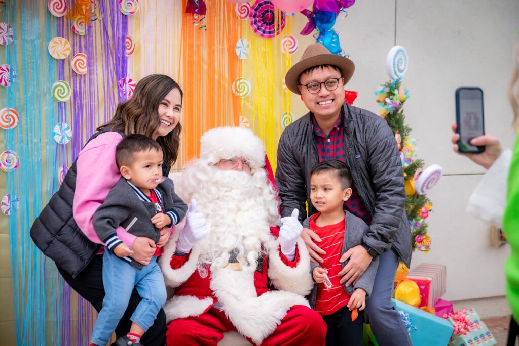 A family with two young children poses for a photo with Santa Claus, who is seated. The background is decorated with vibrant, colorful streamers and lollipops. A small Christmas tree with ornaments and wrapped gifts is visible to the side.