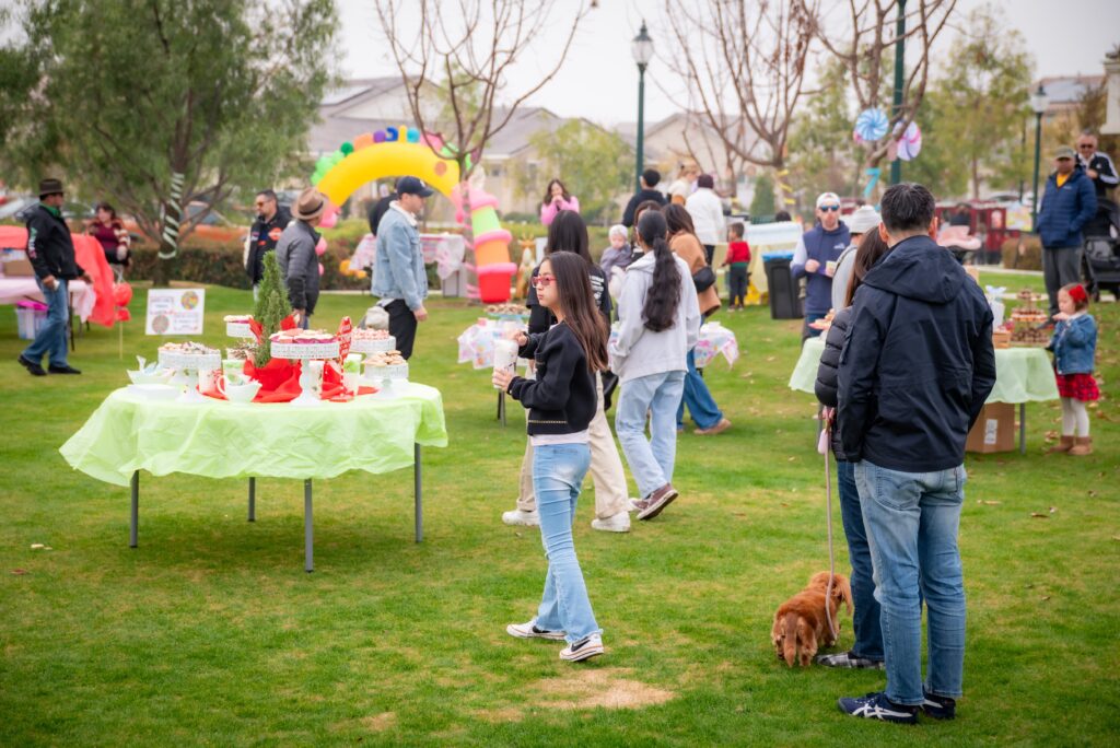 A lively outdoor gathering with people chatting and enjoying a festive atmosphere. Tables with colorful decorations and treats are set up on a green lawn. A person walks a dog, and children play near a colorful inflatable structure.