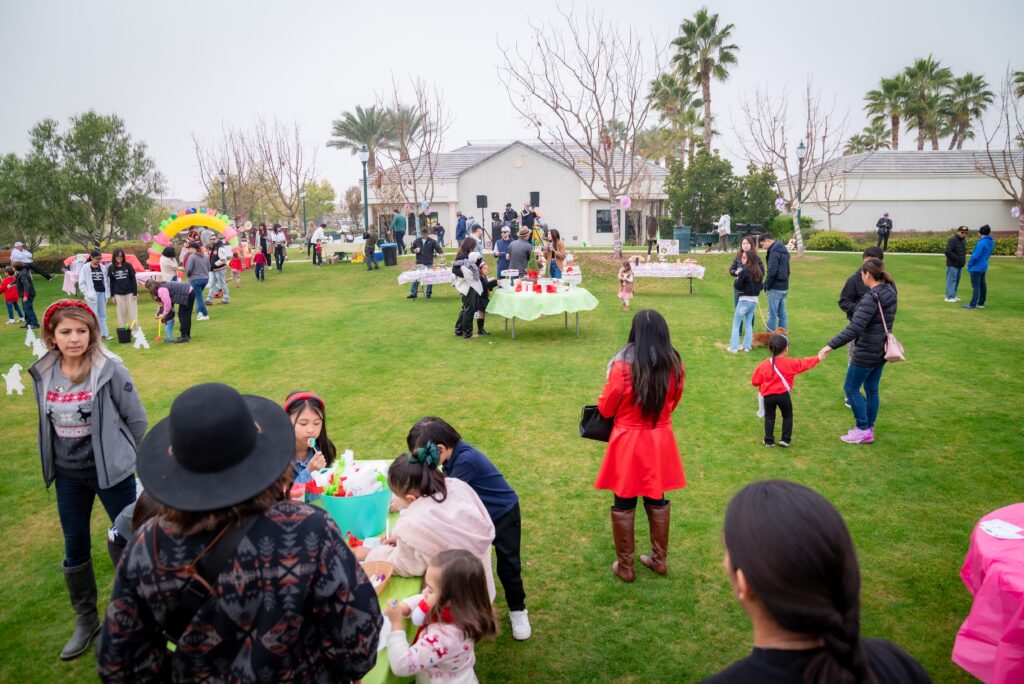 A lively outdoor gathering on a grassy field with families and children engaging in various activities. Kids are playing and adults are socializing near tables covered with bright tablecloths. Trees and houses can be seen in the background.