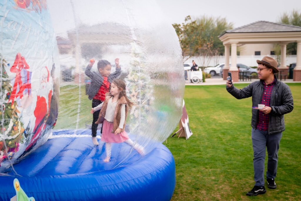 Children play inside a large inflatable snow globe on a grassy area, with holiday decorations visible. A man stands nearby, taking a picture with his phone. Trees and a gazebo are in the background under a cloudy sky.